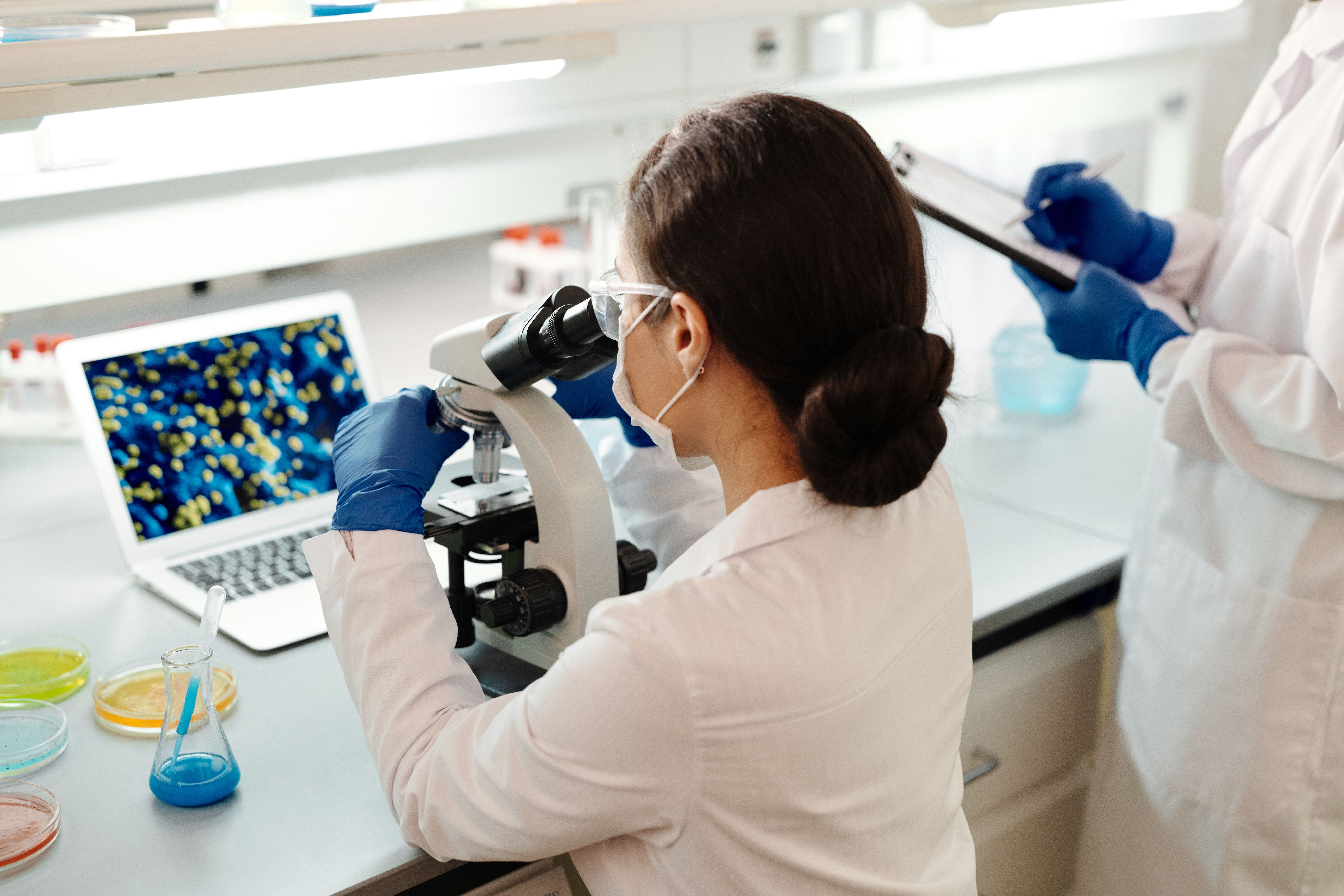 A researcher at a microscope in a clinical lab while a colleague records notes
